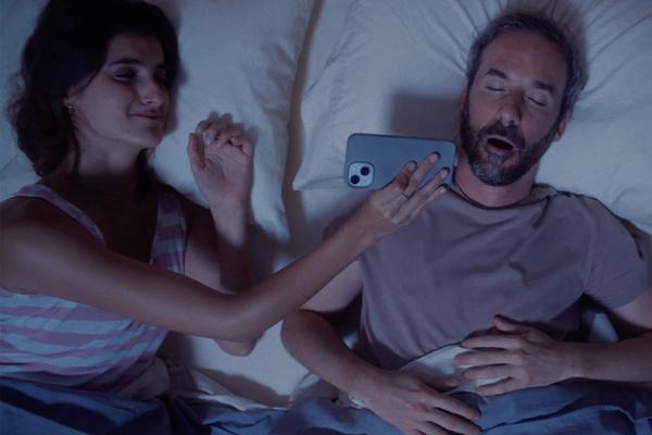 A close-up of a woman in bed smiling while holding a smartphone in front of a sleeping man's open mouth. The man has a beard and is lying on his back.