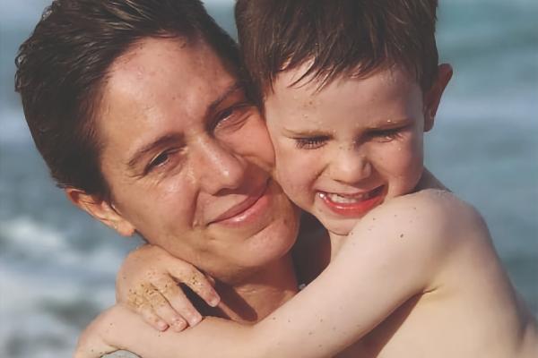 A mother holds her son, both smiling cheek-to-cheek, with the ocean in the background