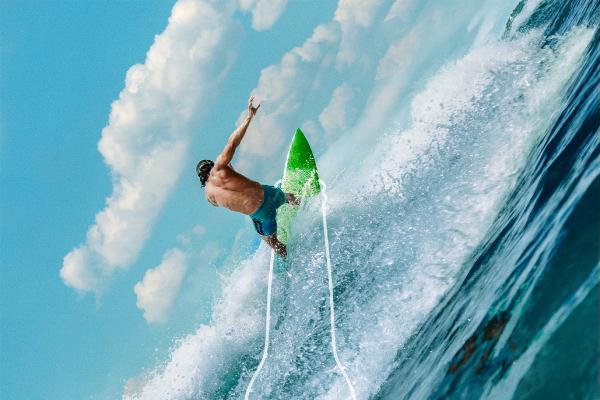A surfer on a green surfboard rides a large blue wave, with a white outline of a beer bottle superimposed, framing the surfer and extending down the wave. The sky is light blue with white clouds.