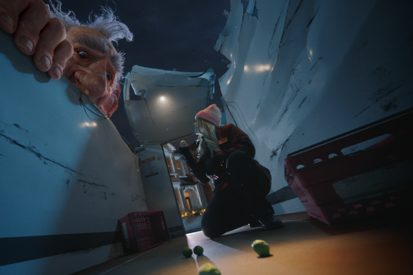 From a low-angle perspective, the animated giant's head and a large hand are seen peering down from above a white surface. Below, a young woman with a pink beanie, dark jacket, and jeans kneels on a dimly lit floor, holding a small object in her hand. Several green, round vegetables are scattered around her on the floor, implying the vast scale difference.