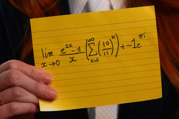 A person's hand holds a yellow lined index card displaying a complex mathematical equation written in black ink. The equation includes a limit as x approaches 0, an infinite summation from k=0 to infinity, and a term with Euler's identity (e to the power of pi i).