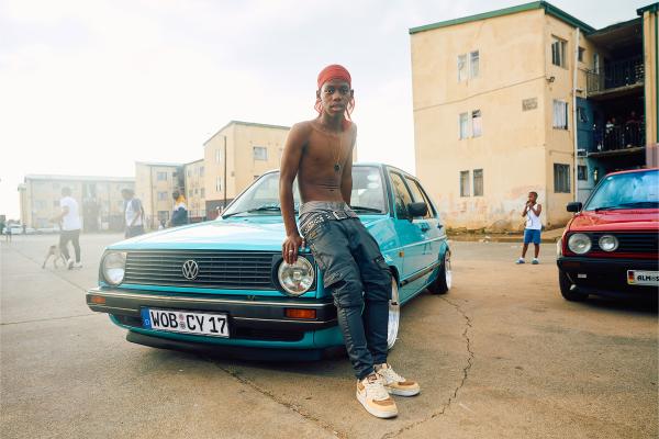 A young, shirtless man with a red head covering and dark pants leans casually on the hood of a teal vintage Volkswagen Golf. He is looking directly at the camera. The car has a German license plate that reads "WOB CV 17". In the background, there are apartment buildings and other people, including another red vintage car on the right. The setting appears to be an urban outdoor area with a light sky.