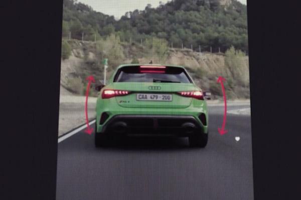A close-up of a smartphone screen displaying the rear view of a bright green Audi RS3 hatchback on a race track. Red curved arrows on either side of the car suggest interactive controls or a visual representation of movement. The car's license plate reads "CAA 479-200". In the background, green trees and hills are visible under a light sky.