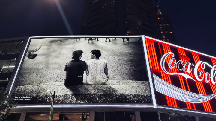 An urban night scene showcasing a large, illuminated billboard featuring a black and white photograph. The photo depicts two people sitting on a concrete ledge, viewed from behind, with the word 'Enjoy' visible on the back of one person's shirt. A pigeon is mid-flight in the upper left of the billboard image. Below the main image, text reads '4:27 pm is Coca-Cola time. @ Kings Cross Rd & Victoria St.' To the right of this billboard, a prominent, vibrant red and white neon sign spells out 'Enjoy Coca-Cola' i