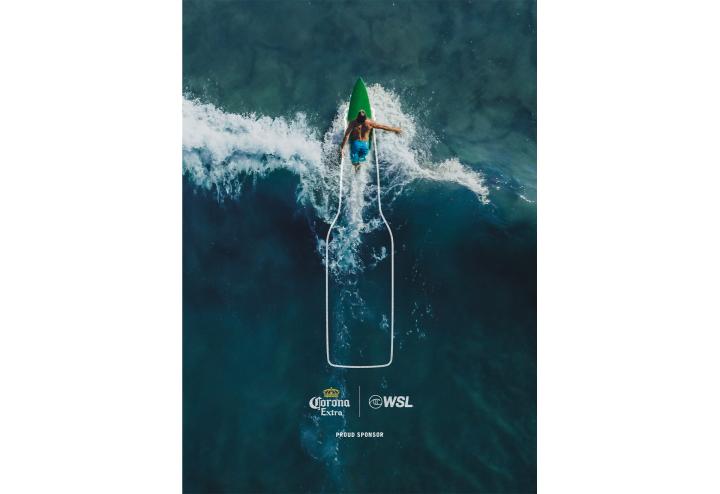 An aerial view of a surfer on a green surfboard riding a wave in dark blue water. A white outline of a beer bottle frames the surfer and extends down the wave. The Corona Extra and WSL logos with "PROUD SPONSOR" text are at the bottom.