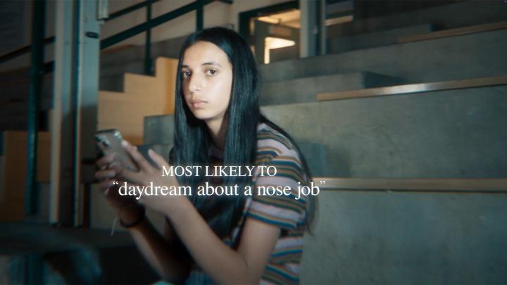 Most Likely To — Dove Young girl sitting on school gym bleachers holding smartphone. White overlay text "Most likely to daydream about a nose job"
