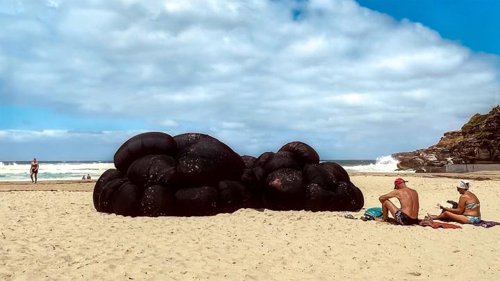 The Spot — Glue Society and The Beautiful and Useful Studio Large fabric art installation on a beach that depicts a melanoma. Two people sit on the beach next to the installation