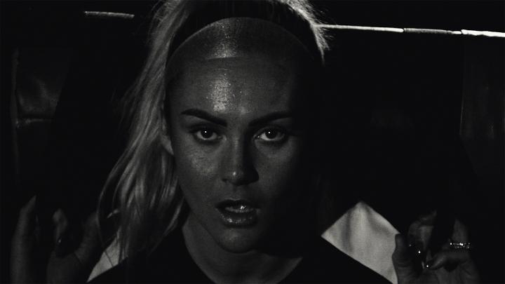 A black and white close-up of a woman's face, her skin appearing sweaty and shiny, with a serious expression and dark background. She has light hair pulled back in a ponytail.