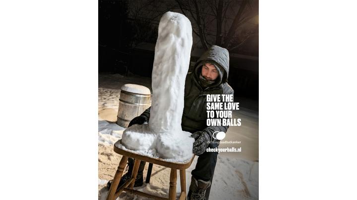 A person in a hooded winter jacket and gloves looks at a tall, phallic snow sculpture perched on a wooden stool in a snowy outdoor scene at night. White text overlaid on the image reads: "GIVE THE SAME LOVE TO YOUR OWN BALLS," followed by "stichting zaadbalkanker" and "checkyourballs.nl".