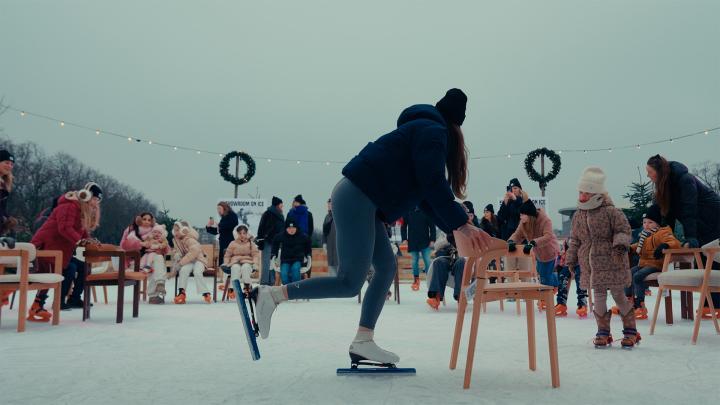 A wide shot shows a woman in a black jacket and hat, wearing ice skates, pushing a light wooden chair across an outdoor ice rink. Many other people, including adults and children, are also on the ice, some learning to skate with chairs or sitting on them, with string lights overhead and signs in the background.