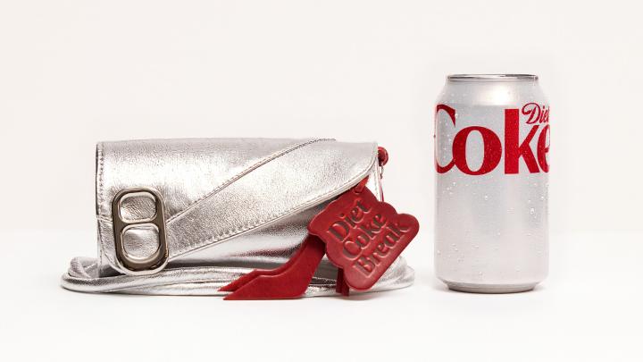 A silver metallic clutch purse with a red "Diet Coke Break" tag sits beside a condensation-covered Diet Coke can on a white background.