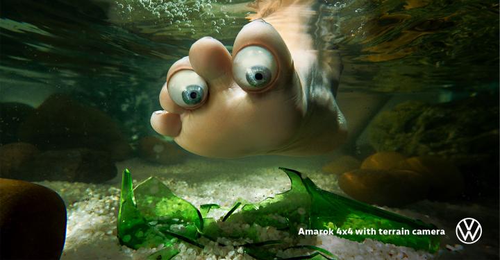 Underwater shot of a foot with eyeball-like toes near broken green glass and pebbles, with a Volkswagen logo in the corner.