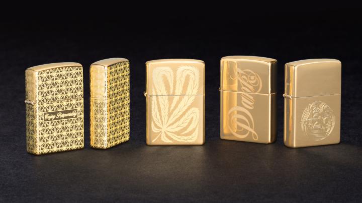 High-Class Collection — Zippo 5 gold Zippo lighters standing upright against a black background. This is the High-class Collection. the first two feature an engraved marijuana leaf pattern, with "enjoy responsibly" engraved in script. The middle one has one large marijuana leaf engraved into it. the 4th has the word "Dank" engraved in script across the face. The far right one has "420" engraved on it in a circular, continuous font and pattern