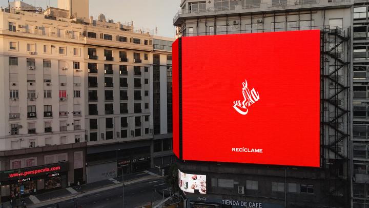 Large outdoor ad in a city. The ad is a red background featuring the logo of Coca-Cola as though it was on a can than had just been crushed. Underneath in white type it says "Reciclaime"