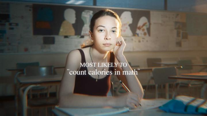 Most Likely To — dove Girl sitting in shcool classroom with hand on chin and holding a pen to a notebook. White overlay text: "most likely to count calories in math"