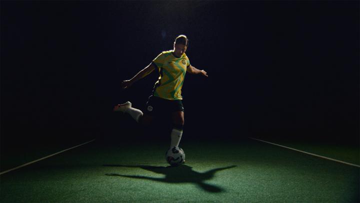 A soccer player in a yellow and green patterned jersey and shorts is kicking a soccer ball, illuminated by a spotlight on a dark green field with white lines. A strong shadow is cast behind them.