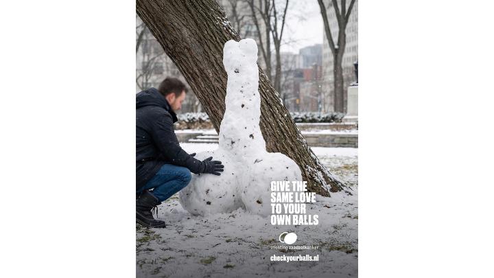 A man in a dark winter coat and gloves kneels on a snow-covered ground next to a large, phallic snow sculpture that is built against the trunk of a tree. White text overlaid on the right side of the image reads: "GIVE THE SAME LOVE TO YOUR OWN BALLS," followed by "stichting zaadbalkanker" and "checkyourballs.nl".