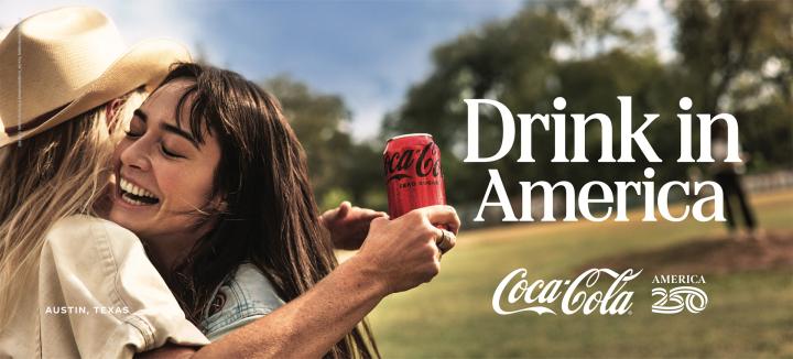 Two young women embrace and smile, with one holding a red Coca-Cola Zero Sugar can. The text "Drink in America" and "Austin, Texas" is visible against a sunny, grassy background.