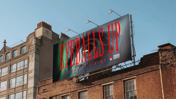 A large billboard advertisement mounted on the side of an old brick building in an urban setting. The billboard features a close-up of a striking human eye, with the words "EYEBALLS UP" in large, red, sans-serif font overlaid across the image. Smaller text "CANADIAN FILM FEST" is visible on the right.