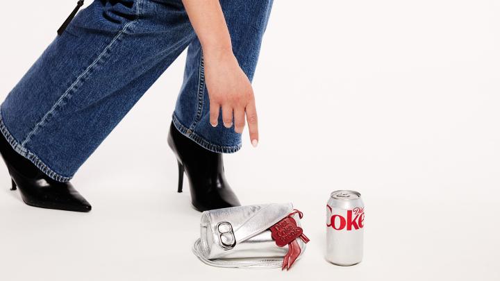 A person in blue jeans and black high heels reaches down towards a silver metallic clutch purse and a Diet Coke can, both resting on a white surface. The purse has a red "Diet Coke Break" tag.