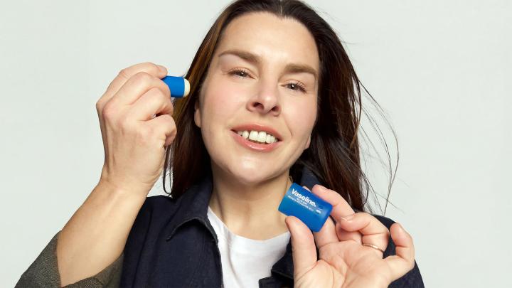 A woman with dark hair smiling gently, holding a blue Vaseline Primer & Highlighter Jelly stick to her cheek and a smaller blue Vaseline square container in her other hand.