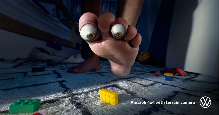 Close-up of a foot with eyeball-like toes stepping on Legos, with a Volkswagen logo in the corner.