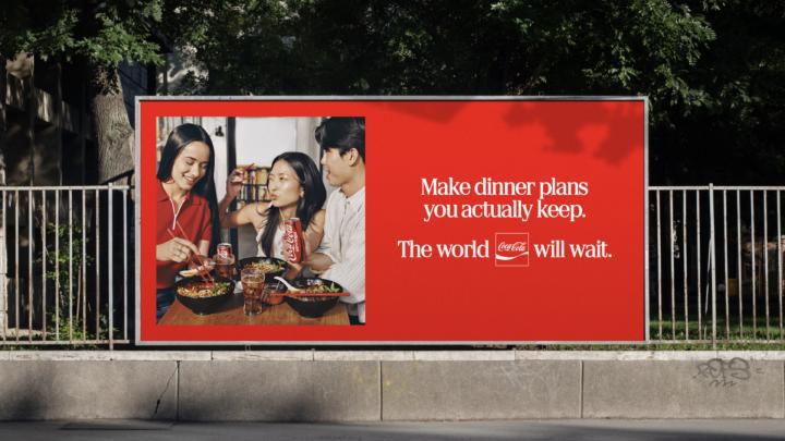 A red Coca-Cola billboard advertisement featuring a photo of three friends sharing a meal on the left, and text on the right reading: "Make dinner plans you actually keep. The world [Coca-Cola logo] will wait."