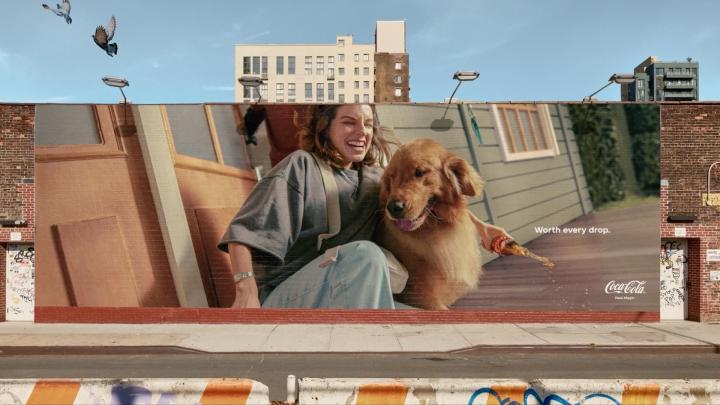 Spills — Coca-Cola Image of a tall outdoor billboard. On the billboard, a woman is playing on a porch with a dog. She is holding a glass bottle of Coca-Cola sideways, and it is spilling out of the bottle.