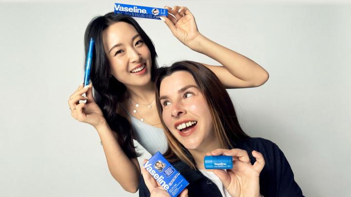 Two women posing together and smiling at the camera, holding various blue Vaseline beauty products. The woman in the back holds a Brow Tamer, and the woman in front holds a Primer & Highlighter stick and a product box.
