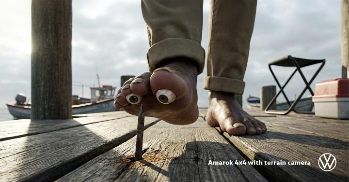 Close-up of a foot with eyeball-like toes stepping on a nail on a wooden dock, with a Volkswagen logo in the corner.