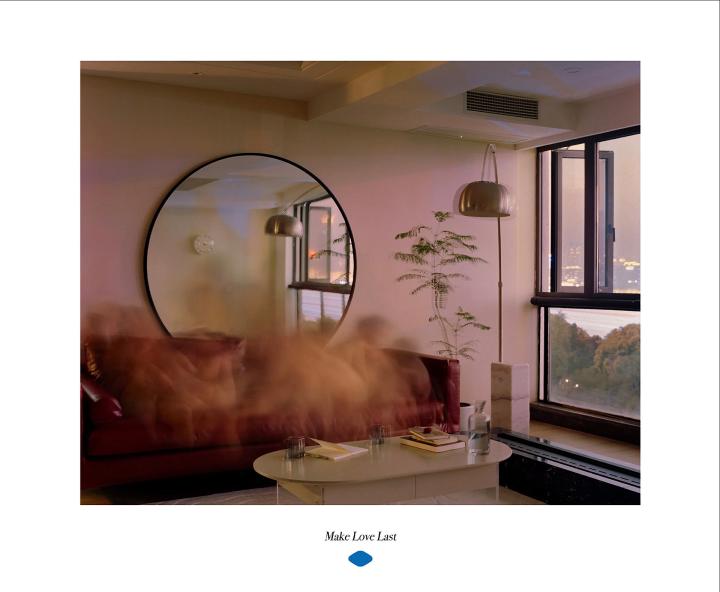 Make Love Last — Viagra A modern living room at dusk, featuring a red sofa, a round mirror, and a view of the city skyline. A coffee table holds glasses and books. A blurred, cloud-like effect obscures the center of the image. The text 'Make Love Last' is displayed below.