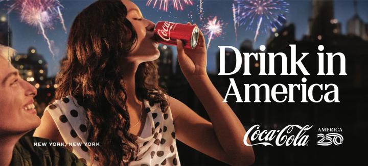 A woman with long dark hair drinks from a red Coca-Cola can, illuminated by city lights and fireworks exploding in the night sky. Another person is smiling to her left. The text "Drink in America" and "New York, New York" is visible.