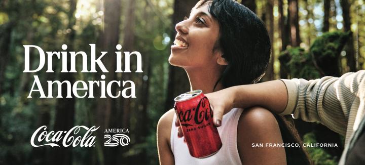 A young woman with dark hair looks upwards, smiling broadly, while holding a red Coca-Cola Zero Sugar can. An arm with a hand rests on her shoulder. The background is a sunny, lush forest. The text "Drink in America" and "San Francisco, California" is visible.