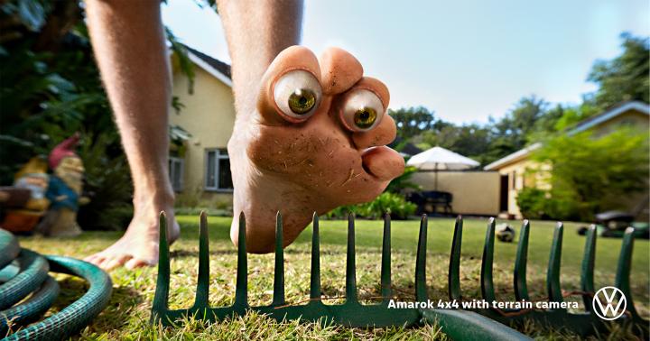 Close-up of a foot with eyeball-like toes stepping over a rake in a grassy yard, with a house in the background.