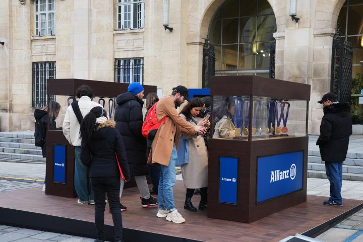 People outdoors at a promotional table with an Allianz banner that is displaying hanging olympic style medals  