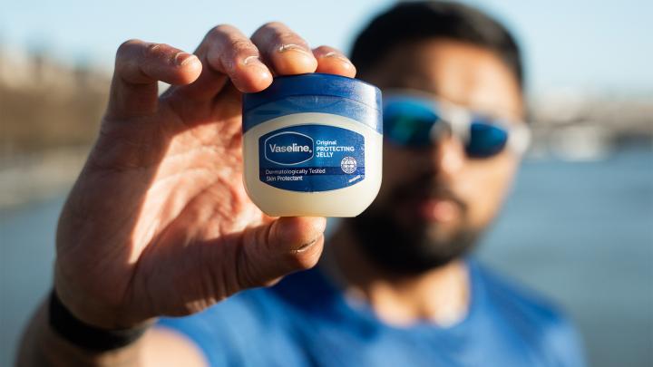 Close-up of a hand holding a Vaseline Original Protecting Jelly jar outdoors, with a blurred runner in the background.