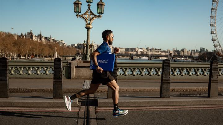 Runner wearing a blue shirt carries a blue Vaseline box while running across a bridge with a city skyline and Ferris wheel in the background.