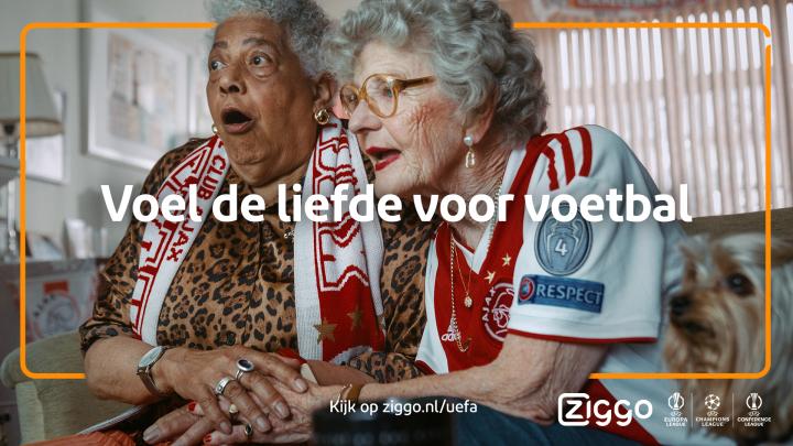 Experience the Love of Football Together — Ziggo Sport Two women sitting on couch with favorite team's gear on watching football match excitedly with text overlay "Voel de liefde voor voetbal"