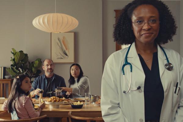 A female doctor with curly dark hair and glasses, wearing a white lab coat and a stethoscope, stands in the foreground looking at the viewer. In the blurred background, a family of three (a man, a woman, and a young girl) is seated around a dining table, engaged in a meal.