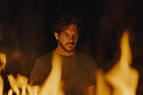 A man with dark hair and a beard, wearing a dark t-shirt, looks intently forward with a serious expression. He is illuminated by the warm glow of flames that are visible in the foreground and background, suggesting he is in a fiery or intense situation.