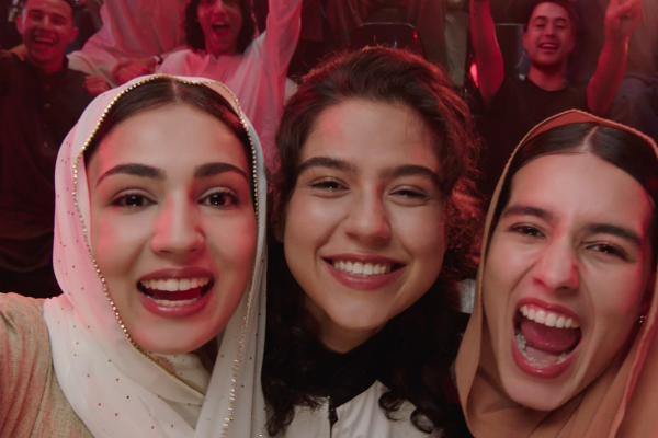 Three young women are captured in a close-up, excitedly smiling and looking directly at the viewer. The woman on the left wears a light-colored headscarf, the one in the middle has dark hair, and the woman on the right also wears a light headscarf, her mouth open as if cheering. In the blurred background, a crowd of diverse people are seen cheering and raising their hands, indicating they are at an event like a concert or sports game.