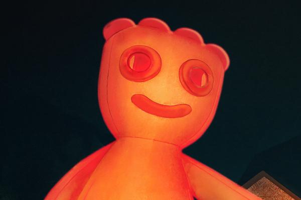A close-up of a large, glowing orange inflatable figure, resembling a Sour Patch Kid, set against a dark background. The figure features round eyes, a smiling mouth, and five bumps on its head, illuminated from within.