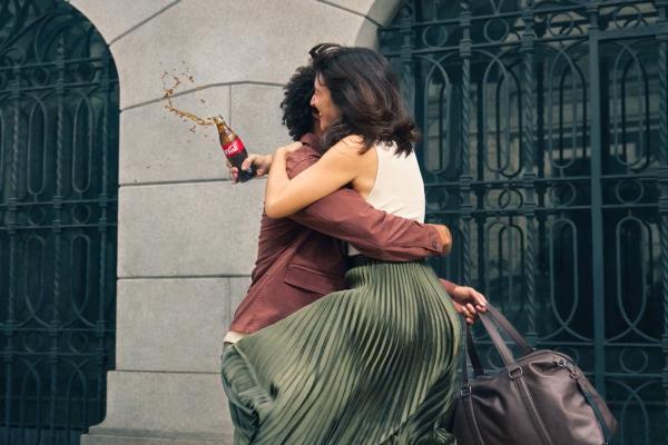 A man and woman embracing. The woman is holding a glass bottle of Coca-Cola which is spilling as she hugs the man