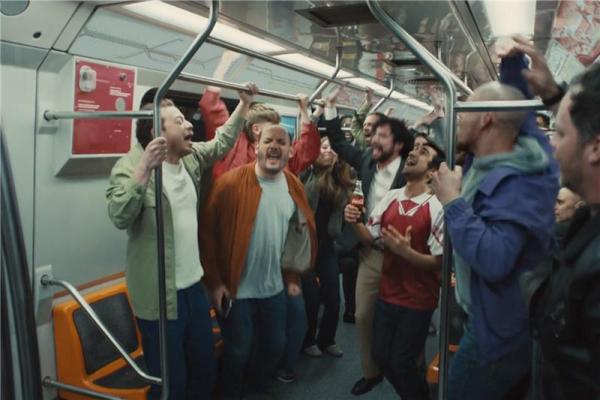 Group of people on a subway train all singing and shouting together. A man in the middle of the group holds a glass bottle of Coca-cola.