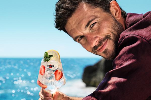 Actor Jonathan Bailey smiling at the camera holding a Martini Spritz with the sea in the background