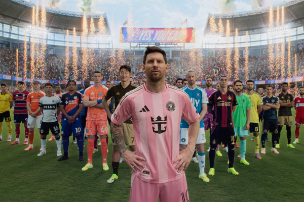 A slightly more cropped, standard-ratio version of the MLS player lineup on the pitch. Lionel Messi stands front and center in his pink Inter Miami jersey with hands on his hips. He is closely flanked by other league players in various colorful club kits. The background features the filled stadium, erupting pyrotechnic sparklers, and the "YES, YOU CAN" video board under a bright, partly cloudy sky.