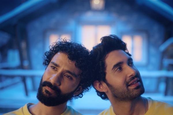 A close-up shot of two men leaning their heads back-to-back, both looking upwards and to the right with expressions of wonder. They have dark hair and beards. The background is softly blurred, showing hints of a snowy, cabin-like setting at night.