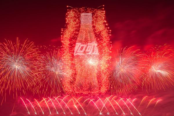 A magnificent fireworks display against a dark red sky. A giant, bright red outline of a Coca-Cola bottle, composed of countless tiny lights or fireworks, prominently features in the center, with Chinese characters inscribed within its shape. Brilliant red and golden fireworks illuminate the sky around the striking bottle.