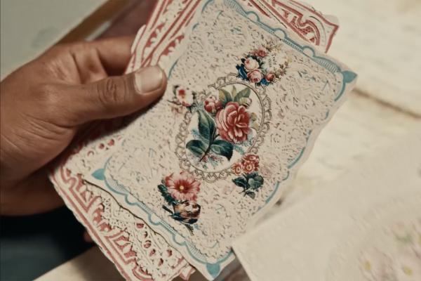 A close-up shot shows a hand holding a stack of intricately designed, vintage-style cards. The top card has delicate, lacy borders in red and blue, and a central oval motif filled with a colorful floral arrangement including red roses, other flowers, and a small bird. The cards appear to be made of paper with cut-out details