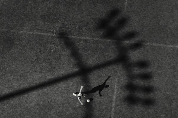 An overhead black and white shot of a person playing soccer, with a ball at their feet and a long shadow stretching behind them on a textured field.